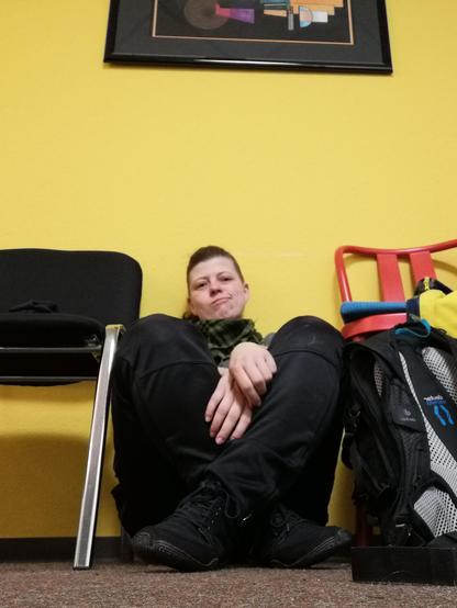 Jaylee sitzt am Boden, an eine gelbe Wand gelehnt. Sie schaut mit leicht skeptischem Blick in die Kamera, die Beine sind aufgestellt und an den Knöcheln überkreuzt. Die Füße stecken in schwarzen Barfussschuhen. Daneben ein roter und ein schwarzer Stuhl. Oben ist noch der Rand eines gerahmten Bildes zu erkennen.