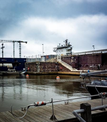 This photograph shows a harbor scene under a cloudy sky at the Brunsbüttel Lock. In the foreground, a wooden jetty with mooring equipment is visible, and to the right, a small boat is partially visible. The water is calm, and several orange and white buoys are floating. In the middle ground, a large brick wall with stairs and a white railing separates the jetty from a higher platform. Beyond the wall, a ship's bridge is visible, indicating a large ship docked nearby. To the left, stacked blue shipping containers bearing the words "UNITAS.com" and two tall cranes are visible. The overall mood is serene.