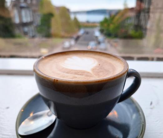 Picture of a Flat White with flowery art standing in front of a window with a blurred view of a street leading away from the viewer, housed to the left and right, and maybe a lake or a river at the bottom (in fact the River Tay)