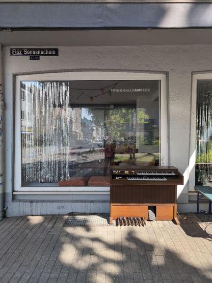 Eine Orgel ist außen vor einem Schaufenster zu sehen. Rechts davon deutet sich eine Sitzbank an. Im Fenster spiegelt sich die Bochumer Straße in Gelsenkirchen.