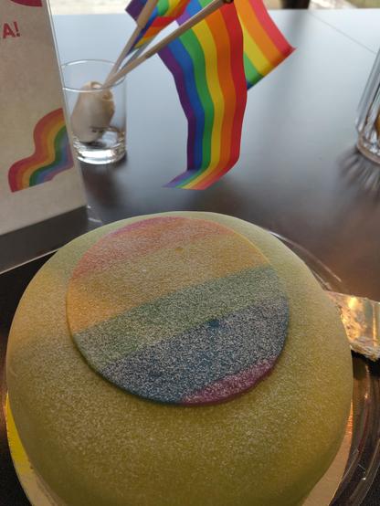 A cake with the pride flag in coloured marzipan, two small pride flags in a glass i the background.