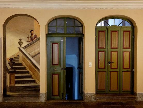 Indoors picture with three symmetric arches. Two contain wooden doors, while the left one contains a trompe l'oeil painting showing a woman descending a grand staircase.