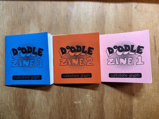 issues 1, 2 & 3 of Doodle Zine sit on a plywood surface. they’re roughly 3x3 inches square and have pink, orange and blue covers respectively.