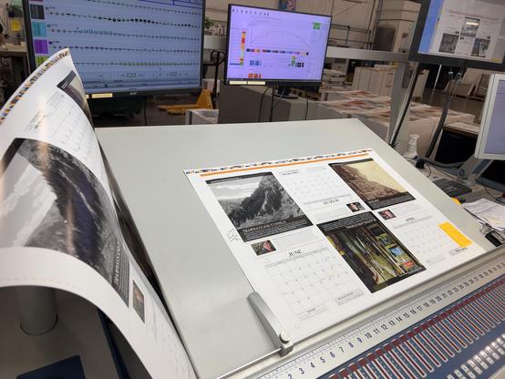A press sheet full of calendar pages I designed, lays on a press man’s proofing table. Various buttons and screens are around it to control the ink coverage and density, etc. 