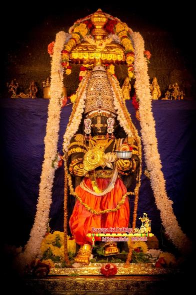 Idol of Hindu deity Krishna in walking pose with the right foot looking slightly ahead of the left. Krishna wears gold shores and radiant red trousers, holds a gold symbol of a disc (chakra) in right hand and a small silver cup in left hand, and wears a stone-studded gold crown. Small gold umbrella over Krishna's head. Thick long white flower garland over the umbrella flowing down either side of Krishna all the way down to the floor. Blue cloth background.