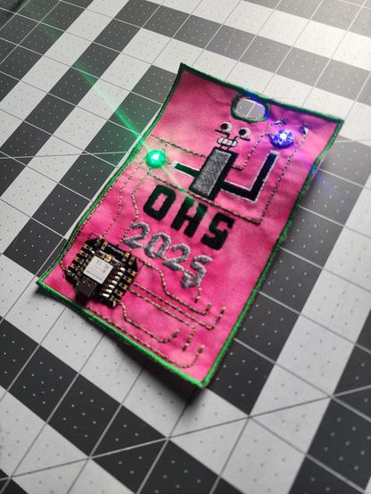 the OHS2025 badge in alternate colour way pink and green with one of the 2025 'little guys' embroidered on it with OHS2025 embroidered below. the badge has an SAO pin and two leds