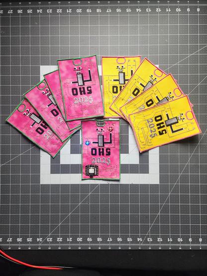 OHS2025 badges fanned out on a table, on the left is the pink and green version and the right the yellow and pink version. in the centre is a pink and green version with an SAO pin and two leds lit up