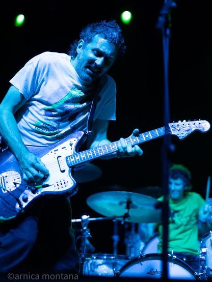 A musician passionately plays an electric guitar on stage, wearing a graphic t-shirt. In the background, a drummer is visible. The stage is illuminated with green lights, creating a vibrant atmosphere.