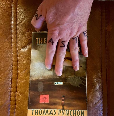 A hand reaching down from the top of the frame to touch a well-worn copy of Thomas Pynchon’s The Crying of Lot 49. On the fingers of the hand are the letters W, A, S, T, & E, written in black Sharpie. 