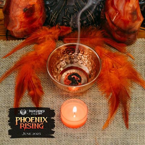 A hammered copper bowl sits on a burlap-covered surface, surrounded by red and orange feathers. There is a substance burning in the bowl, sending up a thin stream of smoke. This is one of the items in the exclusive June 2025 "Phoenix Rising" Inked Goddess Creations Box.