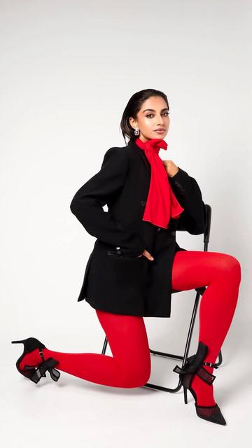 Meenakshi Chaudhary's fashion-forward ensemble includes a black blazer, red leggings, and a dramatic red scarf. The outfit is accessorized with ruby earrings and black stilettos, reflecting her confident and chic style.