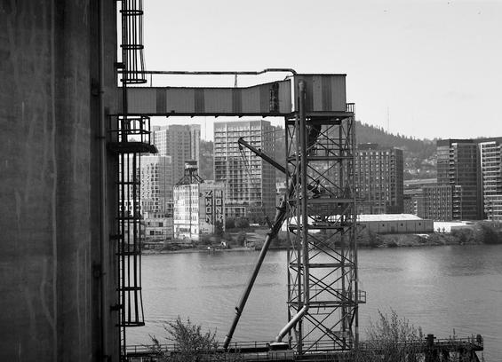A black and white photograph looking west across the Willamette river at the NW Portland skyline. In the foreground is an old grain structure for loading ships and though that structure is a small, old, abandoned building across the river with large graffiti down the side saying "Zwon" and "Monk" the words being possibly 30-40 feet tall. 