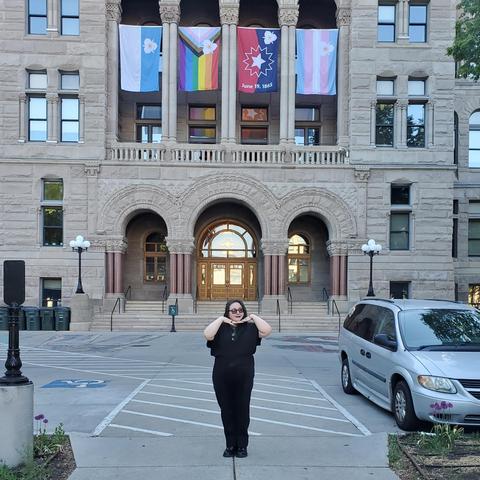 An adorable queerdo standing in front if the SLC City & County Building, which is displaying the new official city flags.

"Each of the new designs maintains the tri-symmetrical Sego Lily symbol that serves as the traditional city flag's canton, but with the blue-and-white background replaced with the imagery of the Juneteenth Celebration, LGBTQIA Belonging and Transgender Visibility flags."