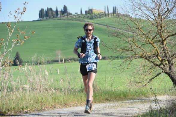A man wearing ultra running gear running on a gravel road with a backdrop of a Tuscan hilltop village