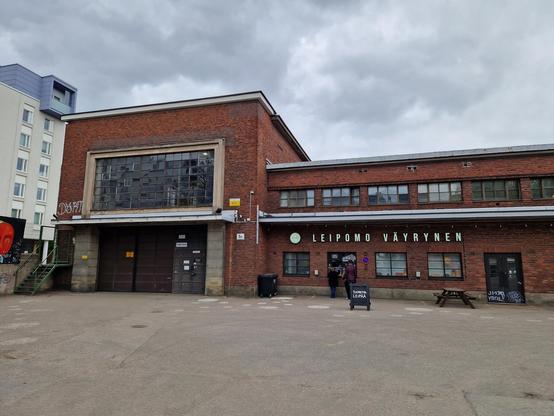 A low industrial building made of brick, with three people queuing at a window, under a sign reading Leipomo Väyrynen (Bakery Väyrynen).