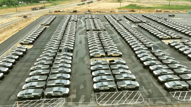 **Fotobeschreibung fuer Sehbehinderte:**

Das Bild zeigt ein riesiges, geordnetes Parkfeld voller identischer Tesla Cybertrucks. Die futuristisch anmutenden Fahrzeuge mit ihrer kantigen, silber-metallischen Karosserie sind in streng symmetrischen Reihen abgestellt – hunderte davon. Die Szene wirkt industriell und auf Effizienz ausgelegt. Der Asphalt ist stellenweise noch nass, vermutlich von einem frischen Regenschauer. Im Hintergrund erkennt man eine Baustelle mit erdigem, aufgerissenem Boden, Baufahrzeugen und vereinzelten Baeumen. Die Umgebung laesst auf ein Werk- oder Logistikgelaende in ländlicher oder halb-urbaner Umgebung schliessen – sehr wahrscheinlich in der Naehe der Tesla Gigafactory Texas.
