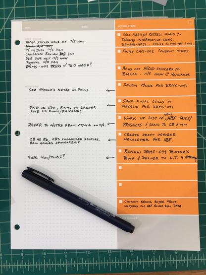 Overhead shot of a notepad on a tabletop. The notepad had an orange column on the right and a larger white column on the left. The orange column has rows of checkboxes with an area next to them to write in to-dos. There are various project related action items written in black ink in the columns. A black pen rests on top of the pad. 
