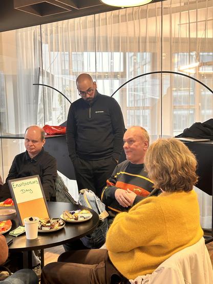  A group of people gathered around a round table at the Envision office, chatting and sharing snacks. A small sign on the table reads “Envisioners Day.”