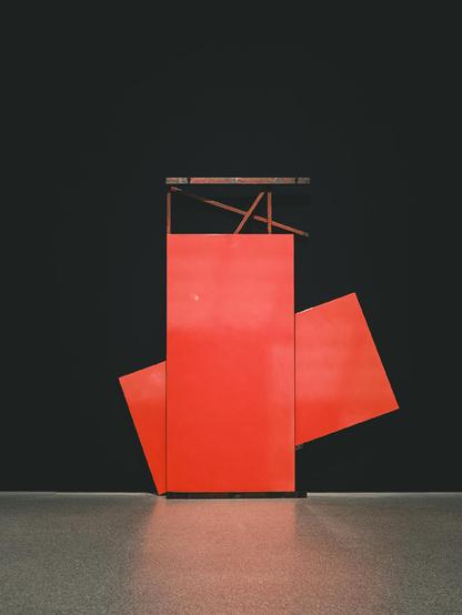 A bold, geometric sculpture made of vivid red panels arranged in an abstract composition, set against a dark background with a polished gray floor. Thin, rusted metal beams intersect above the main red structure, adding contrast and texture.