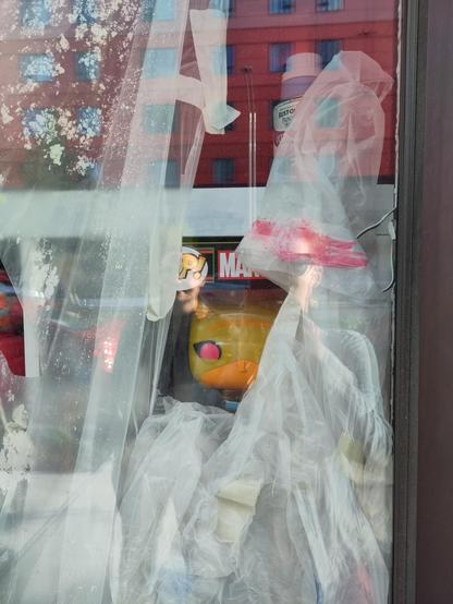 Photo of a storefront window mostly covered with tape and plastic sheeting while the shop undergoes some kind of renovation. A single Funko Pop appears to peer at the viewer from behind a curtain of plastic sheeting. (I don't know what character it's supposed to be.)