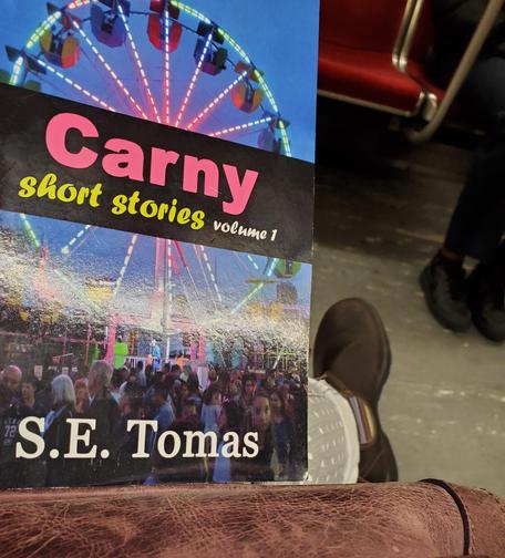 Me holding the book Carny short stories volume 1 while sitting on the subway - my leg glad in gray and white polka dot jeans and foot shod in a brown Blundstone with a colourful side insert are visible in the background.