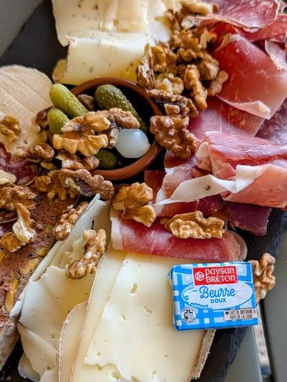 A delectable spread of charcuterie and cheese, featuring various cured meats, an assortment of cheeses, crunchy walnuts, and small pickles, accompanied by a pat of Paysan Breton butter.