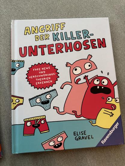 Das Bild zeigt das Buchcover von "Angriff der Killer-Unterhosen" von Elise Gravel. Das Cover ist in hellen Farben gestaltet und zeigt mehrere cartoonartige Unterhosen mit Gesichtern, die in verschiedenen Farben wie Rot, Blau und Gelb sind. Die Unterhosen haben unterschiedliche Muster und Ausdrücke, einige erscheinen erschrocken oder ängstlich. Im Zentrum des Covers sind drei große, farbenfrohe Monsterfiguren, die erschrocken wirken. Der Titel des Buches ist in großen, bunten Buchstaben geschrieben: "ANGRIFF DER KILLER-UNTERHOSEN". Darunter steht in einem roten Stern: "FAKE NEWS UND VERSCHWÖRUNGSTHEORIEN ERKENNEN". Der Name der Autorin, Elise Gravel, steht unten auf dem Cover, sowie das Logo des Verlags Ravensburger. Das Buchcover liegt auf einem grauen Textil, vermutlich einem Tisch oder einem Sofa.