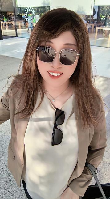 Me smiling while taking a selfie outside the Apple Park Visitor Center. I was wearing sunglasses, a beige blazer over a cream blouse, and had long light brown hair. The Apple logo and glass building facade are visible in the background.
