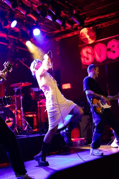 A live music performance featuring a singer in a white dress and fishnet stockings, energetically singing into a microphone. Two musicians are playing electric guitars beside her. The stage has colorful lighting and a visible drum set in the background.
