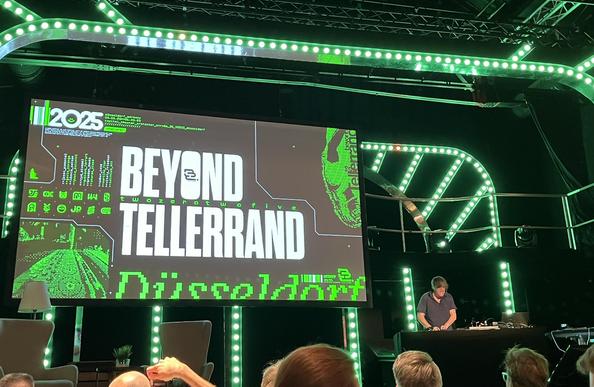 A green-lit stage with a large screen on which “beyond tellerrand Düsseldorf” is written in white and green colors. On the right-hand side, a DJ can be seen standing at a console.