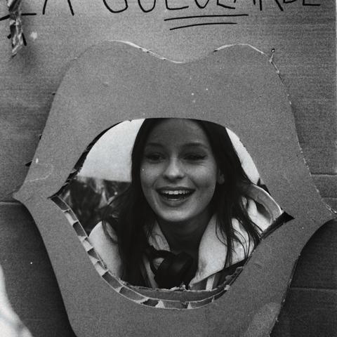 Portrait d'une jeune femme souriante dont le visage apparaît au centre d'un panneau en carton découpé en forme de bouche, lors d’une manifestation. Autour d’elle, des pancartes féministes. La scène évoque la parole libérée et la puissance douce de l’expression engagée.