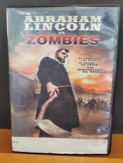 Photo of the front of a dented and scuffed DVD case on a wooden library shelf. It shows an illustration of Abraham Lincoln holding a wooden-handled, rusty scythe in his right hand, looking down at that viewer, from an angle which may indicate the view of a recently dispatched zombie. Lincoln wears a black long-sleeved wool coat, and has his trademark beard and brown hair. In the background is a cloudy sky illuminated orange with what looks like fire from wooden buildings of a city. In the middle distance, a horde of zombies approaches.

Text reads: Abraham Lincoln (white serif old-time block letters) Vs. (small white sans-serif block letters) Zombies (red sans-serif block letters with a gradient color of red at the top to black at the bottom). Sub text  at the center-right reads "He emancipated the slaves... He saved the Union... And slaughtered the Undead!" (black serif block letters). There is a solid white strip insert at the bottom of the illustration which is part of the library's system of labeling the spines of the DVD cases.