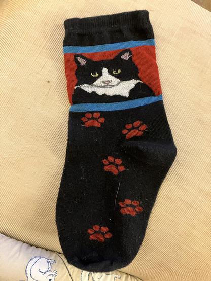 A black sock featuring a design of a tuxedo cat's face on the upper part, with red and blue stripes. The lower part of the sock has several red paw prints.