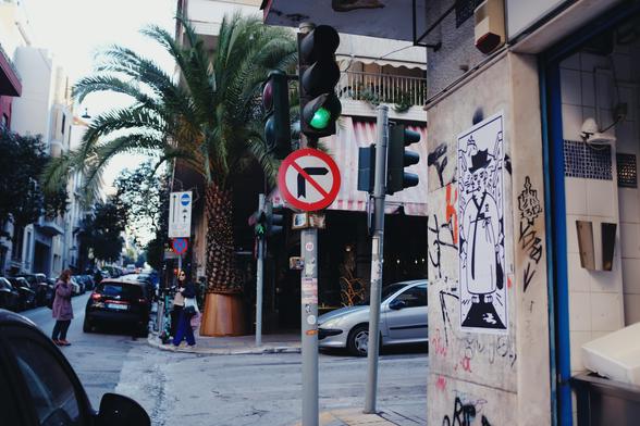 An urban street scene on Kallidromiou Street in Exarcheia, Athens, featuring a no right turn sign, traffic lights, a palm tree, parked cars, pedestrians, and a corner shop wall with street art, specifically a drawing of a tiger walking through the farmers market.