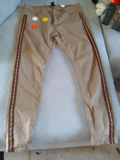 Un pantalon marron clair avec des galons noir, blanc, jaune, orange et blanc sur les côtés. Sous la poche droite il y a une fleur blanche, une jaune et une orange ainsi que le mot "Peace" écrit en lettre dorée.