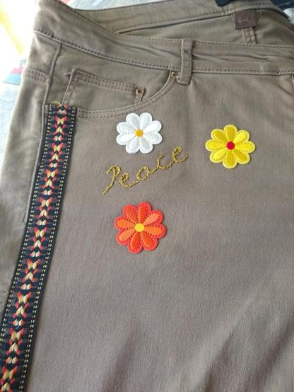 Détail du pantalon précédent. On voit en gros plan les trois fleurs et la broderie. La fleur blanche est au dessus de la broderie qui est écrite en montant. La fleur jaune est au bout de la broderie et la fleur orange est en dessous.