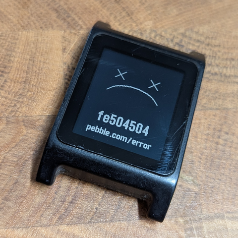 An old Pebble 2 watch without its straps and with a sad face and a fe504504 error message on the screen.
