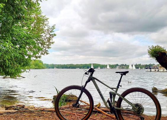 Ein Mountainbike steht an einem Seeufer. Im Hintergrund sins Segelboote zu sehen. 