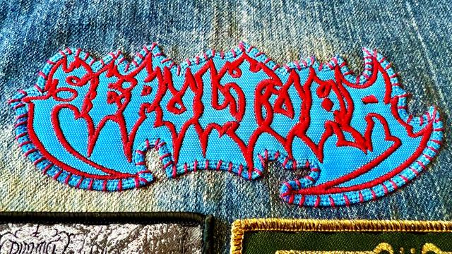 A blue Sepultura patch, whip stitched with red thread, on the lower back of a denim battle jacket. 