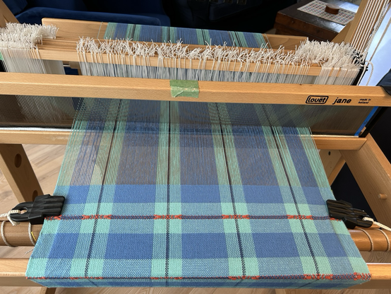 A hand loom with a work in progress on it - a blue checked tea towel with the selveges held by clip temples