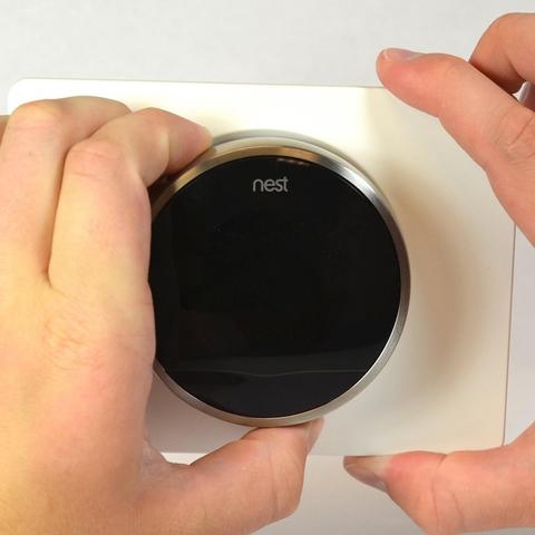 POV image of someone removing or installing a Google Nest thermostat on a wall. Their left hand is holding the thermostat itself, while their right is holding the wall plate to which it's attached.