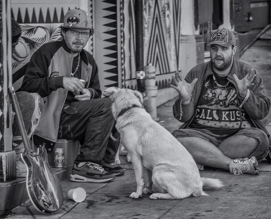 Two young men sit on the sidewalk with a dog. It appears as if one man is telling a story to the dog.