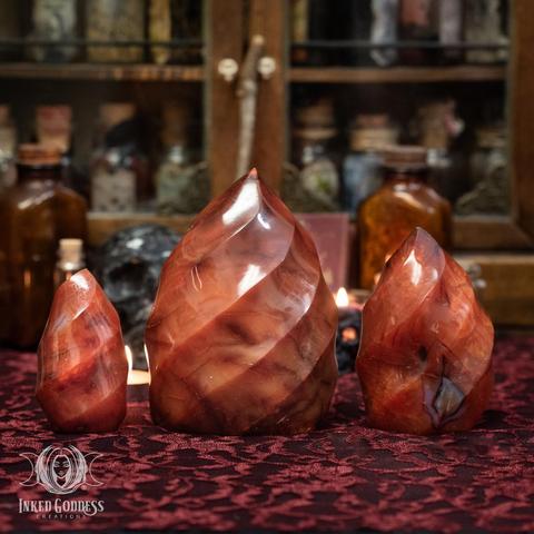 Three specimens of the "Carnelian Flame" product are arranged on a red lace-covered surface. They are carved into a "flame" shape, a chunky form with a flat bottom and carved in spiraling grooves that are reminiscent of fire. Presented by Inked Goddess Creations.