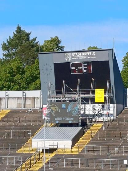 Das Bild zeigt den Stadionplatz von Stadt Krefeld, ein Fußballstadion in Deutschland. Im Vordergrund sind die leeren Tribünen zu sehen, die in dunkelbraunen Sitzreihen angeordnet sind, mit gelben Stufen, die zu den oberen Sitzbereichen führen. Auf der rechten Seite des Bildes ist ein großer, grauer Scoreboard zu sehen, auf dem die Worte "STADT KREFELD" und "Kreativ - Innovativ - Willkommener" stehen. Darunter sind die aktuellen Spielstand und die Zeit angezeigt, wobei der Gastteam mit 14 zu 0 führt. Links neben dem Scoreboard ist ein Banner mit der Aufschrift "Ravens vs Griffirs" zu sehen. Im Hintergrund sind Bäume und ein klarer blauer Himmel zu erkennen. Der Stadionplatz scheint in einem guten Zustand zu sein, mit einigen Baugerüsten, die auf Renovierungsarbeiten hinweisen