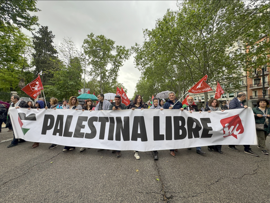 Pancarta principal de Izquierda Unida por el Paseo del Prado. La pancarta blanca dice "PALESTINA LIBRE" en letras negras grandes, con el logo de IU y una logo de una bandera palestina en forma de corazón. Los manifestantes sostienen banderas rojas de IU y banderas palestina. Se ven árboles y edificios a ambos lados de la calle. El día parece nublado. La imagen captura un momento de una protesta o manifestación a favor de la liberación de Palestina.
