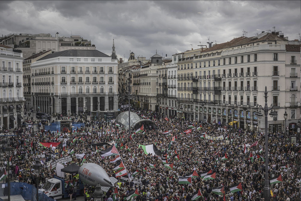 Una vista aérea de una gran manifestación en la plaza de Sol en Madrid.

La plaza está rodeada de edificios clásicos de varios pisos. Una multitud enorme llena la plaza, ondeando muchas banderas palestinas. Se puede ver un gran globo aerostático con la frase "Fin al comercio de armas" en él. El cielo está nublado. La imagen transmite una atmósfera de protesta y activismo político.