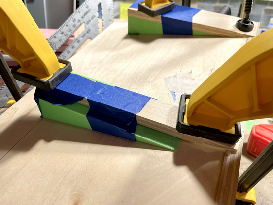 Two slim pieces of wood taped to two neon green 3D printed wedges. The slim wood pieces are being held to a long flat piece of wood with clamps as glue dries