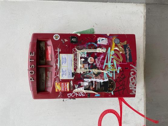 A heavily stickered and graffiti-covered red Italian mailbox standing out against a plain white wall.
