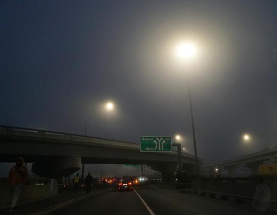A dark, wide view of the sweep of a highway interchange on a misty morning. Street lights tower over the scene like floating orbs, and the red tail lights of cars can be seen retreating in the middle-ground. Shadows of pedestrians walk along the left side.
