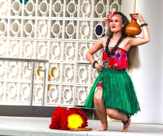 A polynesian dancer from Tupua Productions wearing traditional garb.  The young woman is wearing a green grass skirt and red top with purple flowers  She is squatting down and holding a shiny brown items on her shoulder.